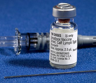 Close-up photo of three components of a smallpox vaccination kit with the vial of vaccine in the foreground on a blue background