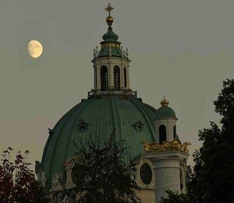A grand domed building with classical architecture, featuring a green copper dome topped by a cupola and cross, stands partially obscured by trees. Ornate golden embellishments and statues accent the structure, especially on a turret to the right. A nearly full moon glows in the muted gray sky, suggesting dusk or dawn, adding a serene contrast to the building’s opulence.