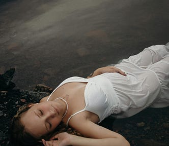 A woman in a white dress lies peacefully at the edge of dark water, eyes closed, symbolizing stillness, reflection, and emotional healing.