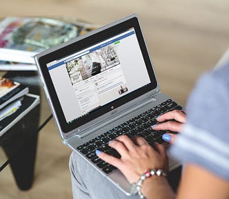 An individual at their laptop looking at a Facebook webpage. Magazines on a coffee table in the background.