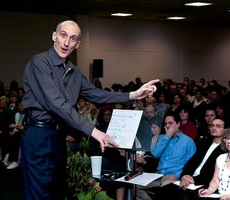 The author, József Manhertz, gives a lecture on a stage in front of an audience of 1,200 people, turns back, and looks into the camera of the photographer standing behind him while speaking.