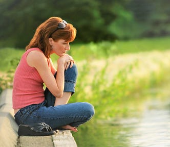 girl with red hair wearing orange top and blue jeans sitting cross-legged on bank of stream with hands under her chin