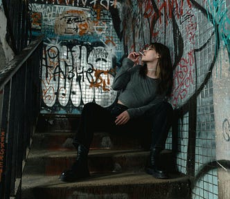 A young woman sits alone on a dimly lit, graffiti-covered staircase, smoking a cigarette. She wears a gray sweater, black pants, and heavy boots, looking off to the side with a distant expression. The setting is gritty, with worn concrete steps and walls covered in colorful, chaotic graffiti, evoking a sense of isolation and despair.