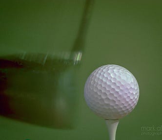 A golf ball on a tee, being hit by a golf club