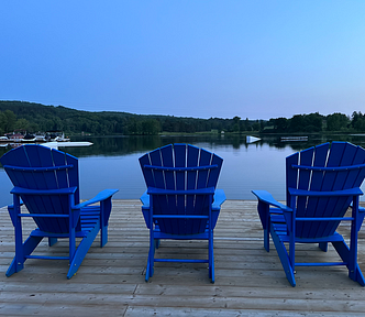 Three blue Muskoka chairs on a dock, facing the lake at dusk.