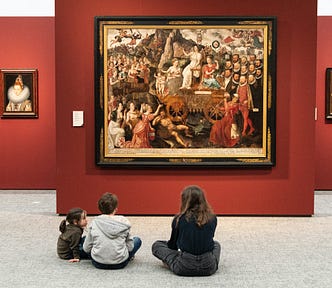 An adult and two small children sitting on the floor of a museum looking up at Tudor-era paintings hung on a deep red wall.