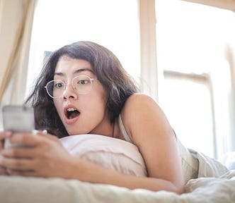 A young woman wearing glasses, lying on a bed, looking at her phone with a shocked expression.