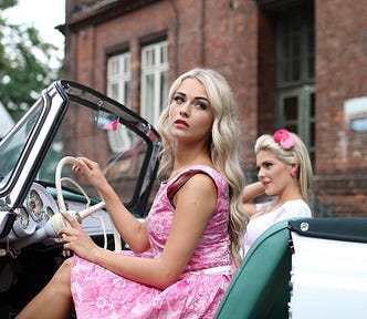 Two beautiful blonde women sitting in the front of a fifties vintage car. The woman driving is wearing a pink sundress. Her passenger has a pink bow in her hair.