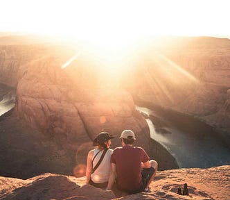 A couple sitting on a rocky ledge in a canyon, quietly watching the sun set over the horizon.