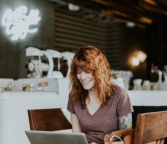 a woman with red hairs smiling and looking at her laptop in a coffe shop