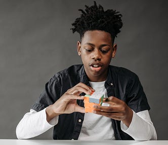 A black boy with locks pointing up above his head as he focuses on a rubiks cube he is holding in his two hands. He is sitting down and wearing a black button up shirt with a white long sleeve below it