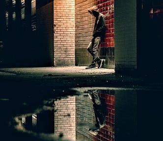 young man wearing hooded sweatshirt leaning against brick wall at night with reflection in puddle