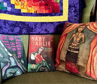 Photo taken by author, of two colorful paperback books sitting on a sage green stuffed chair, next to a red pillow with the likeness of Frida Kahlo, with a rainbow quilt hanging above. The books are called Baby Darlin’ and Laughing In Her Sleep, written by the author’s friends, Alycia and Jon Vreeland, illustrated by Alycia. Jon died in 2020, and Baby Darlin’ is Alycia’s debut novel.