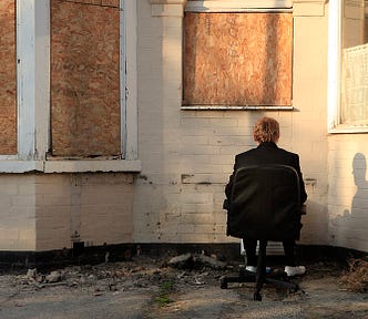 A man sitting in a chair staring at a building that is boarded up