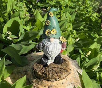 A garden gnome with a green hat decorated with yellow flowers stands atop a tree stump, surrounded by vibrant greenery. The gnome has a long white beard, black boots, and holds a small watering can in one hand and a flower pot in the other. Sunlight highlights the lush foliage and the intricate details of the gnome’s features.