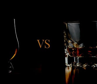 A Glencairn glass with liquor on the left hand side and a tumbler with liquor on the right hand side with the letters ‘VS’ between them against a black background