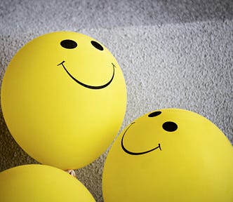 Photo of yellow balloons, against a rough, off-white background. Two and a half balloons are visible in the photo. The two full balloons have smiling faces on them, and the faces are looking at each other.