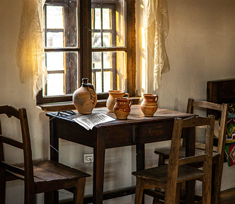 Photograph of a room with golden light streaming in through a wooden framed, 4-pane window with wispy golden-white curtains each side of it. There’s a small table pushed up to the wall directly under the window. It has pottery mugs and a hand drawn cell phone (plus power cord and plug) and papers on it, with three chairs placed at each of the remaining open sides. Part of a wooden bed is in the background on the right, draped in deeply coloured, geometrically patterned fabric.