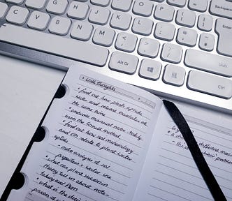 Small notebook with to do list written on it in black pen, sitting next to a white and silver keyboard on a white desk.