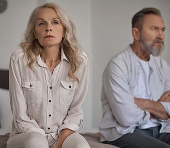 A divided mature couple sit on either side of bed with disappointed looks.