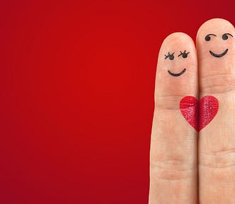 Two fingers close together, the shorter one has female eyes and mouth drawn on it, the taller has male eyes and a smile drawn on it, Each has half a red heart which — side by side — make a whole heart