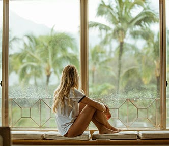 A side view of a young woman with long, blonde hair sitting on a ledge in front of a large window. She is wearing a white t-shirt, but her tanned legs are bare. She is sitting with her bare feet on the seat, and her knees up. The bottom of her t-shirt is gathered on her hips, revealing enough skin to suggest she may be wearing nothing under the t-shirt. Her head is turned away from the camera to look out of the window. The view through the window is of palm trees in heavy rain: hot, and wet.