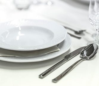 Image shows a background of white, with white plates, silver cutlery and glasses — a set table.