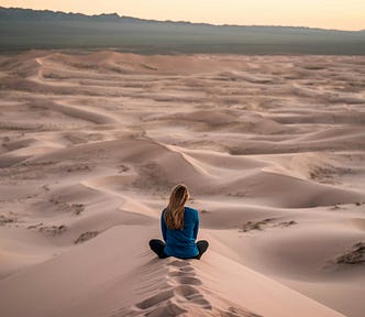 Woman sitting alone in a vast, sandy landscape at sunset, reflecting in stillness beneath a soft golden sky.