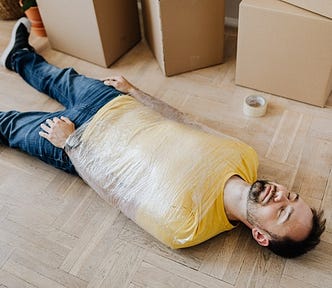 man lying on light wood floor with plastic wrap around his upper torso, boxes in background. exhaustion, adrenal fatigue, adrenals, recharge, thank you.