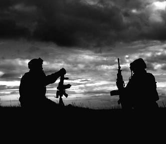 Two soldiers with guns sitting in a black and white photo as silhouettes agains the sunset.