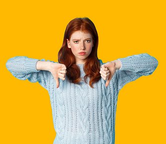 Red haired young woman in a light blue jumper with her arms raised in front of her, with her two thumbs pointing down. She looks a bit negative. Bright yellow background