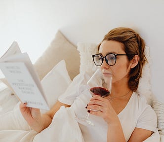 Woman in White Shirt Holding a Glass of Wine reading a book in bed.