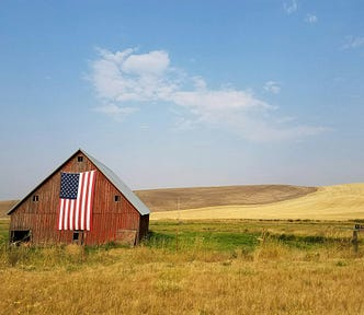 A barn in a very rural area with an American flag draping down from it.