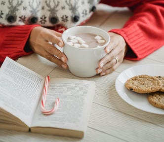 A person drinking a hot chocolate while reading a book. There is a candy cane on the table alongside a plate of cookies.