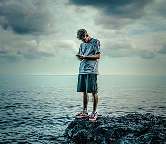 moody picture of young man wearing t-shirt and shorts standing on rock with ocean behind while looking at his phone