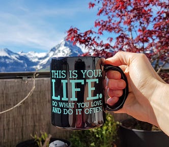 A black coffee mug with a slogan in blue letters: This is your life. Do what you love and do it often. Behind the cup stand a snowed peak and a red-leaf tree.
