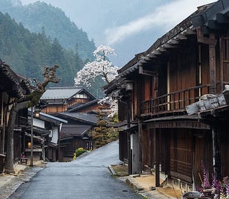 A scenic view of Tsumago-juku, a historic post town on the Nakasendo, during spring. Traditional Edo period (1603–1868) wooden buildings line the quiet street, with a cherry blossom tree in full bloom in the background. Misty mountains and lush greenery enhance the nostalgic atmosphere of this well-preserved town.