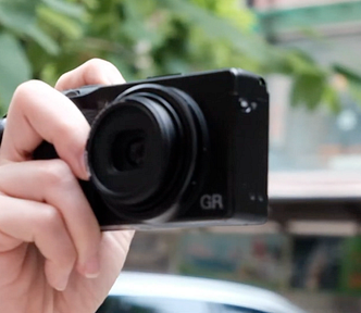 A hand holding a black GR IIIx digital camera, pointing towards the viewer with a blurred background of green foliage and urban streetscape