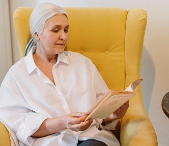 An older woman with hair pulled back sits in a yellow chair reading a booklet. Orange tulips sit on the table beside her, along with a glass of water.