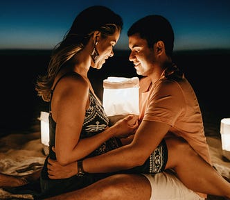 Photo of a man and woman on a blanket on the beach, at dusk. They’re surrounded by portable lights. They’re sitting facing each other, with her legs over him. His arms are holding her waist. Her hands are in between them, holding a white bag filled with white light. They’re both looking down at the light.