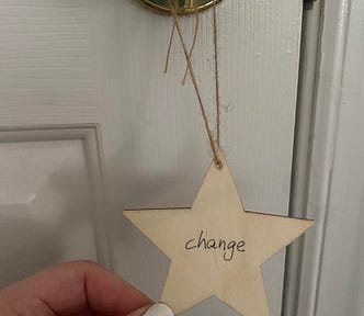 A wooden star ornament with the word “change” is hanging on a gold doorknob. The author’s hand is holding it as it hangs.