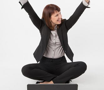 Woman in work attire — suit, and blouse — sits cross legged in front of a laptop and puts both arms in the air to celebrate