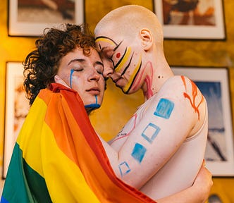 Photo of two women, one light-skinned with a shaved head and the other with slightly darker skin with curly auburn hair. They’re embracing, appearing about to kiss, with a rainbow Pride flag around them, with colorful face and body paint on their face, back, and arm. They’re standing in front of paintings against a yellow wall.