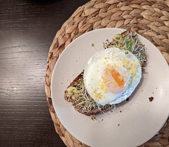 Avocado toast on a white plate, on a wicker placemat, on a black wood table. The avocado toast has a medium egg, sprouts, lemon zest, and everything but the bagel seasoning.