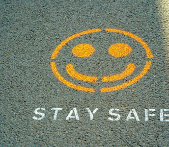 Stencilled road markings, presumably unofficial, on tarmac: a yellow smiley face above white lettering which reads, “STAY SAFE.”