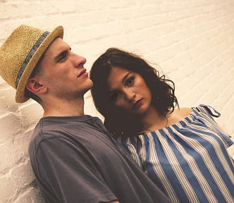 A man and woman stand together against a white brick wall