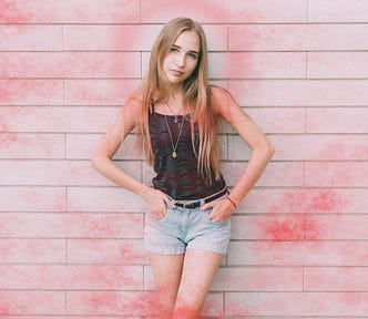 A skinny, blonde girl in her early teens, wearing a dark green tank top and denim cut-offs, leans against a white brick wall. She has the fingers of both hands tucked into the front pockets of her shorts. A red mist obscures much of the scene but her face is clearly visible. She is looking at the camera with an expression somewhere between contempt and wary scepticism.