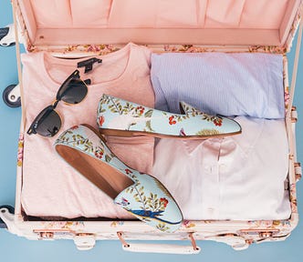 A pink rollerbag carryon suitcase, full of folded clothes in pastel colours, topped by a pair of sunglasses and some white flat ladies’ shoes decorated with flowers