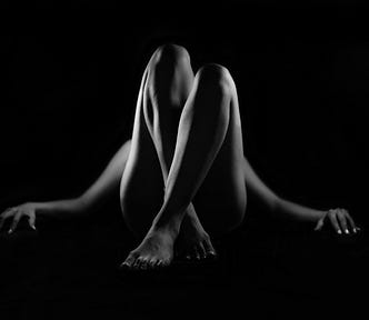 Woman lying down with her legs crossed, knees in air. Viewpoint is looking at the front of the shins. Arms are stretched out to side. Black background with black and white image.
