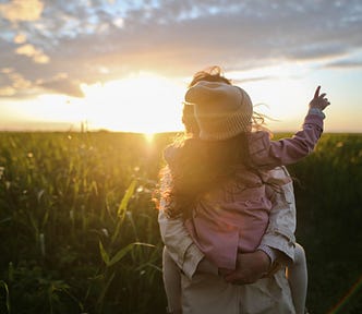 In a grassy field against a setting sun, a woman holds a little girl who is pointing at the sky. They are facing each other and we don’t see their faces.